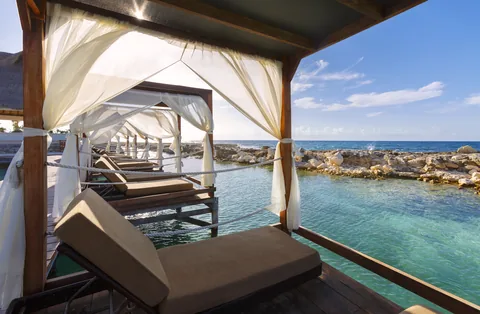 A curtained daybed cabana on a deck above turquoise water.